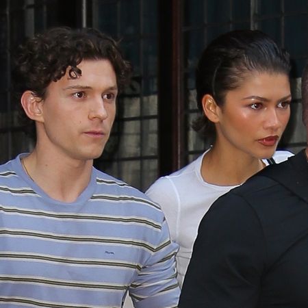 Zendaya wears a white T-shirt and linen pants and has slicked back short hair while holding hands with Tom Holland