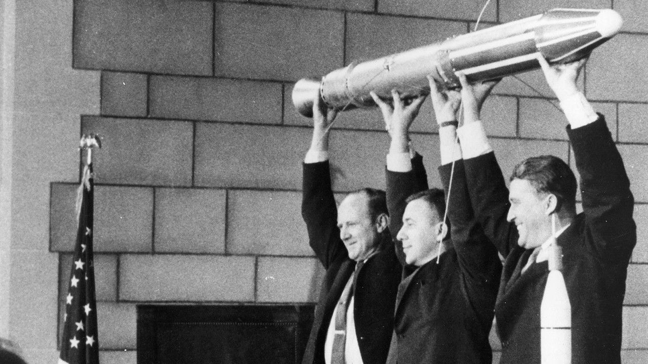 Three men holding a tiny rocket-shaped satellite