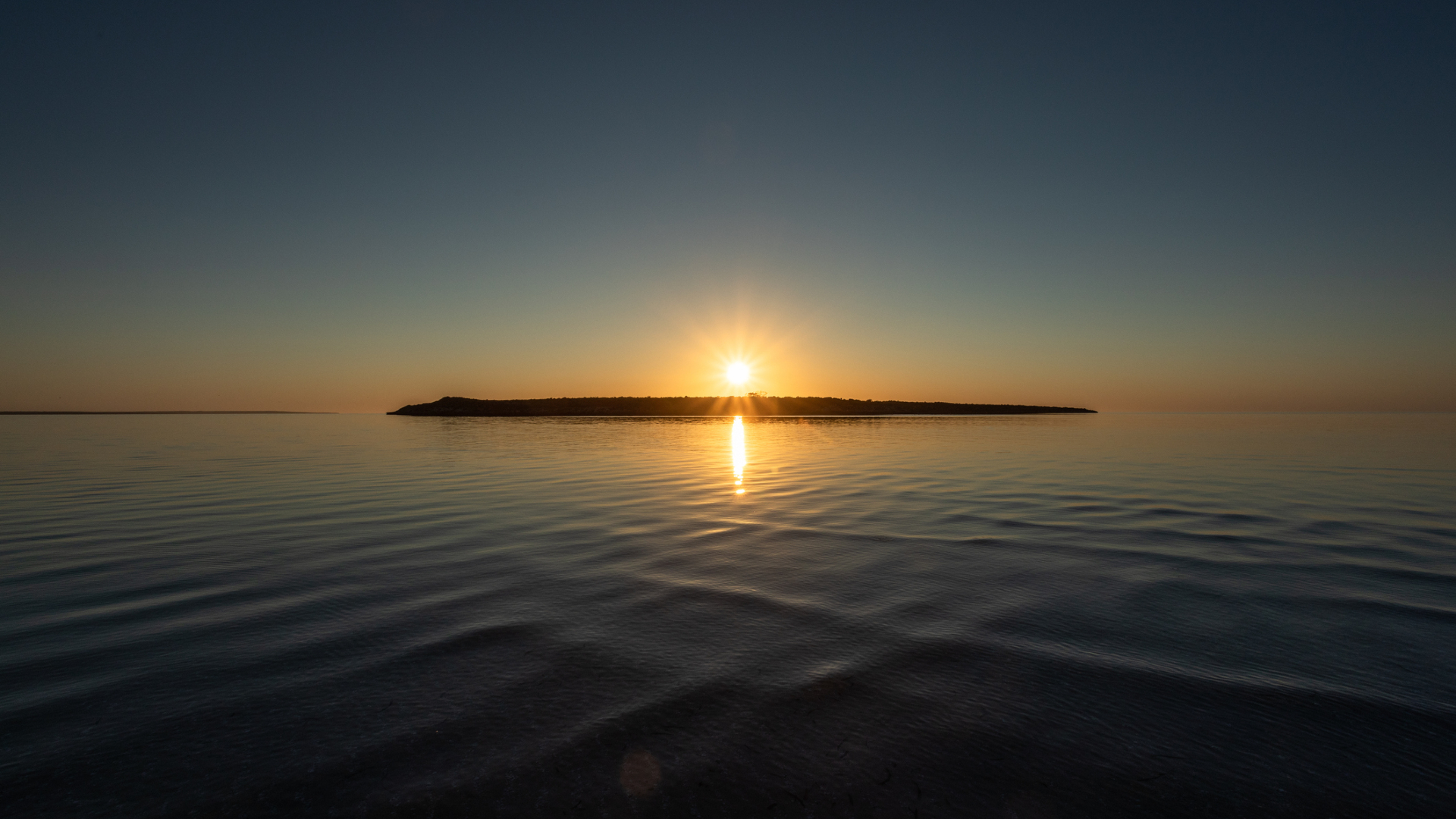 A sunset over a lake with a silhouette of an island right underneath the setting sun
