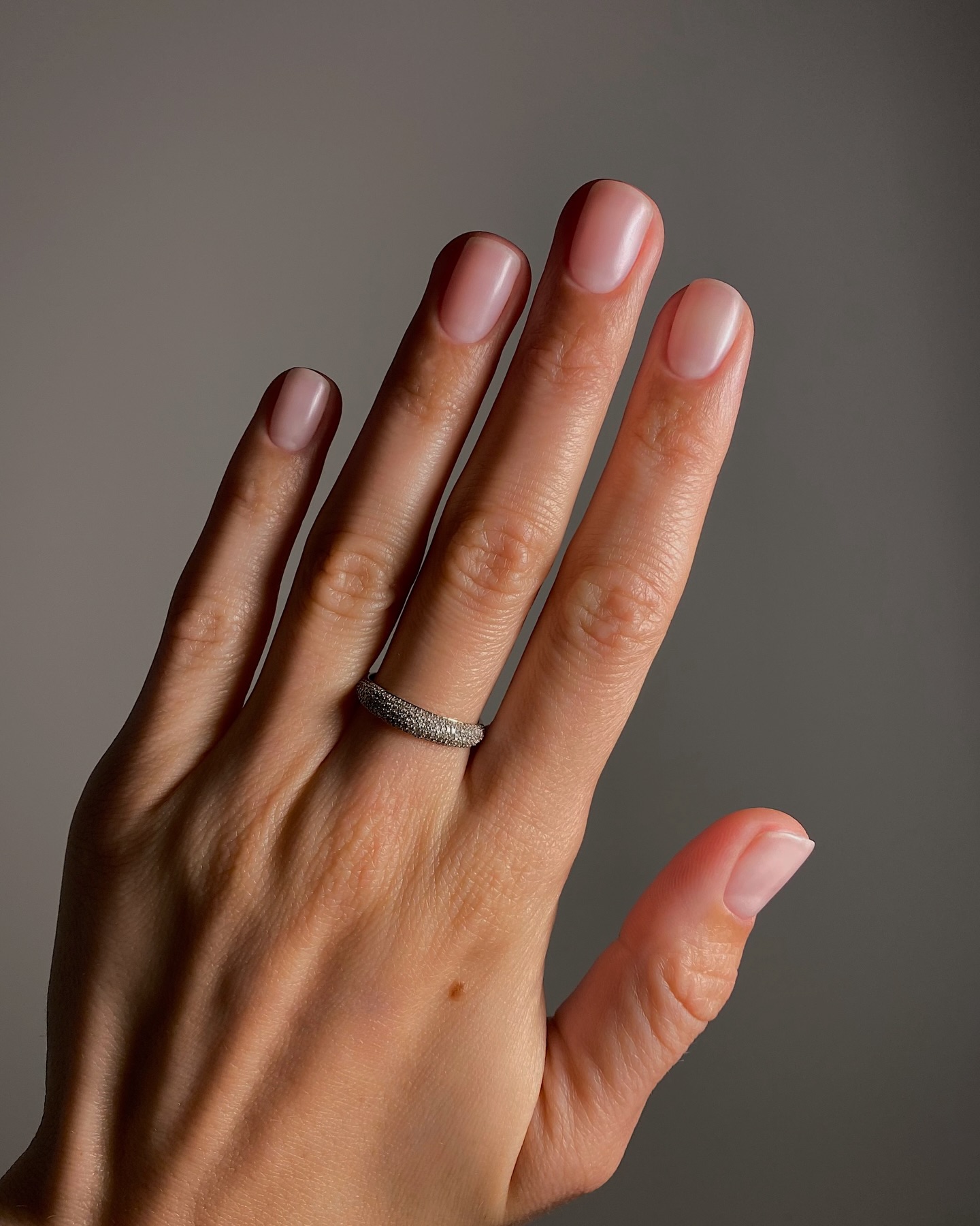 a hand held up in front of a grey-black background and a diamond ring on the middle finger with matter fingernails