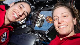 two women astronauts smiling in front of a cupola window. in back is a spacecraft with solar wings, backdropped by earth
