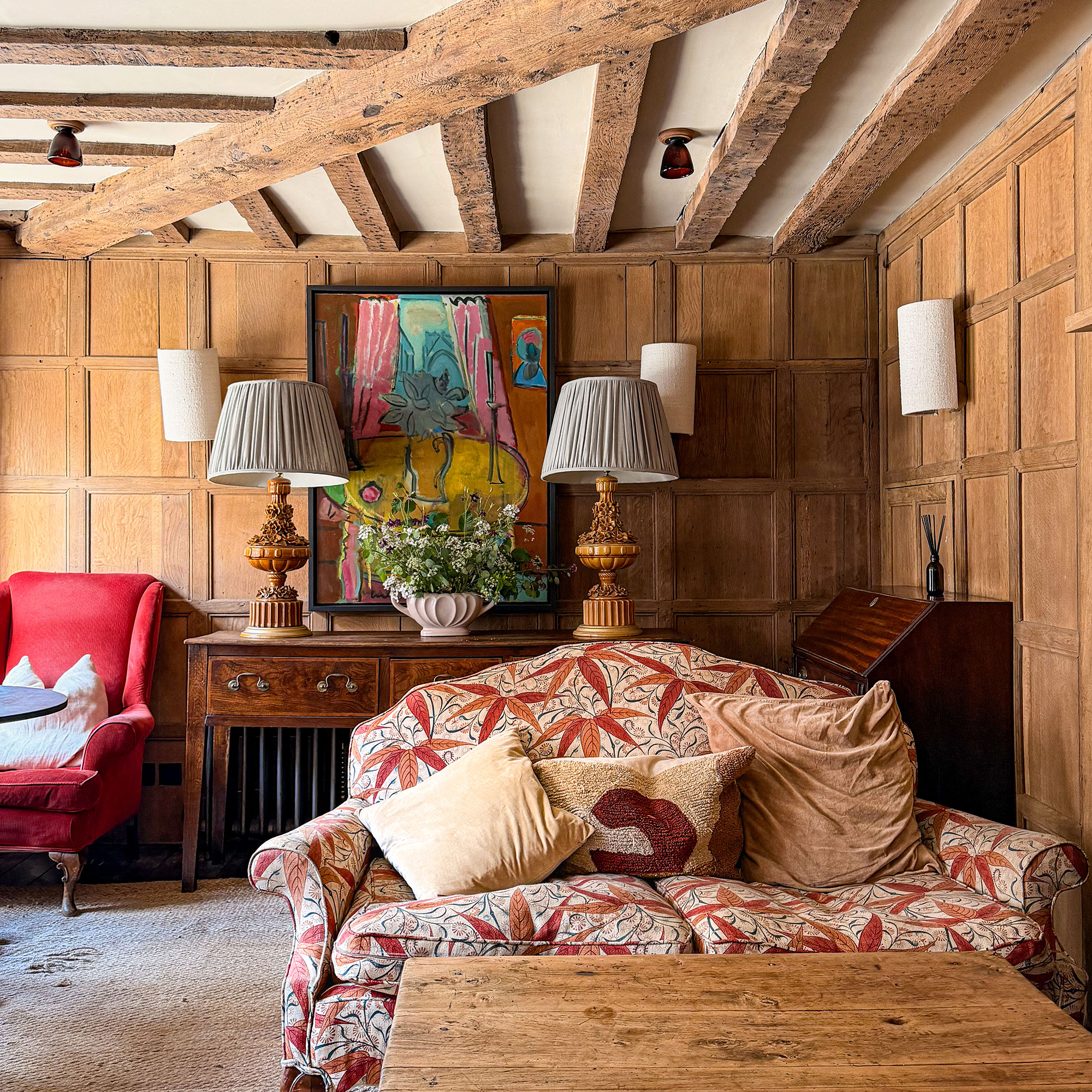Room with wood panelled walls and wooden ceiling beams, red and orange patterned love seat, wooden coffee table and sideboard with vase of flowers and two table lamps