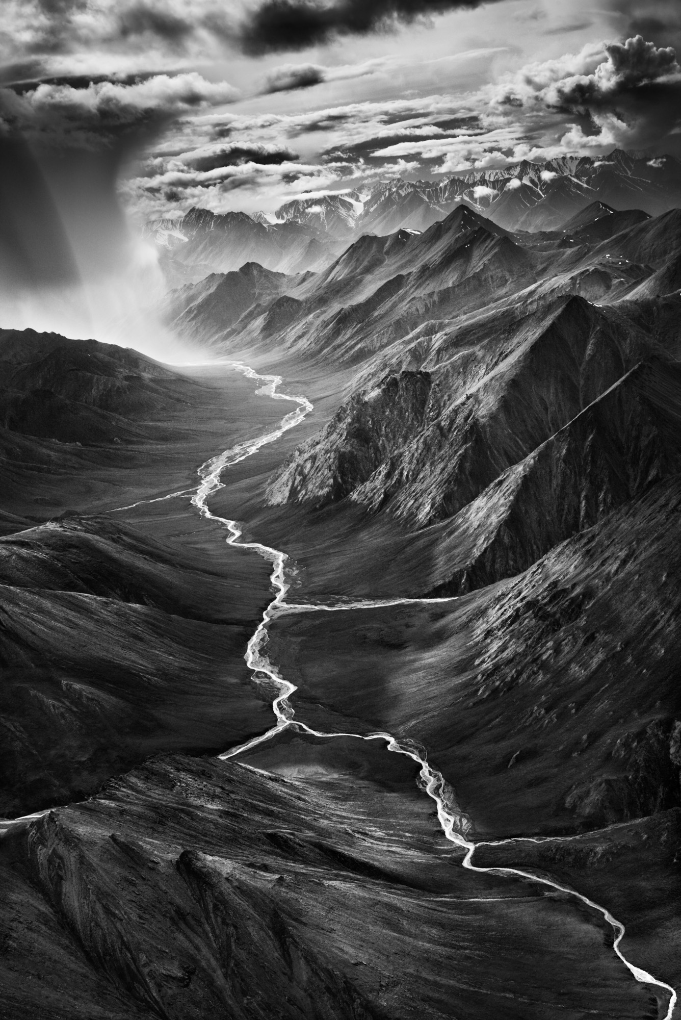 Black and white aerial photograph of Alaska's Brooks Range, a braided river winding through dark mountain ridges beneath a dramatic cloudy sky.