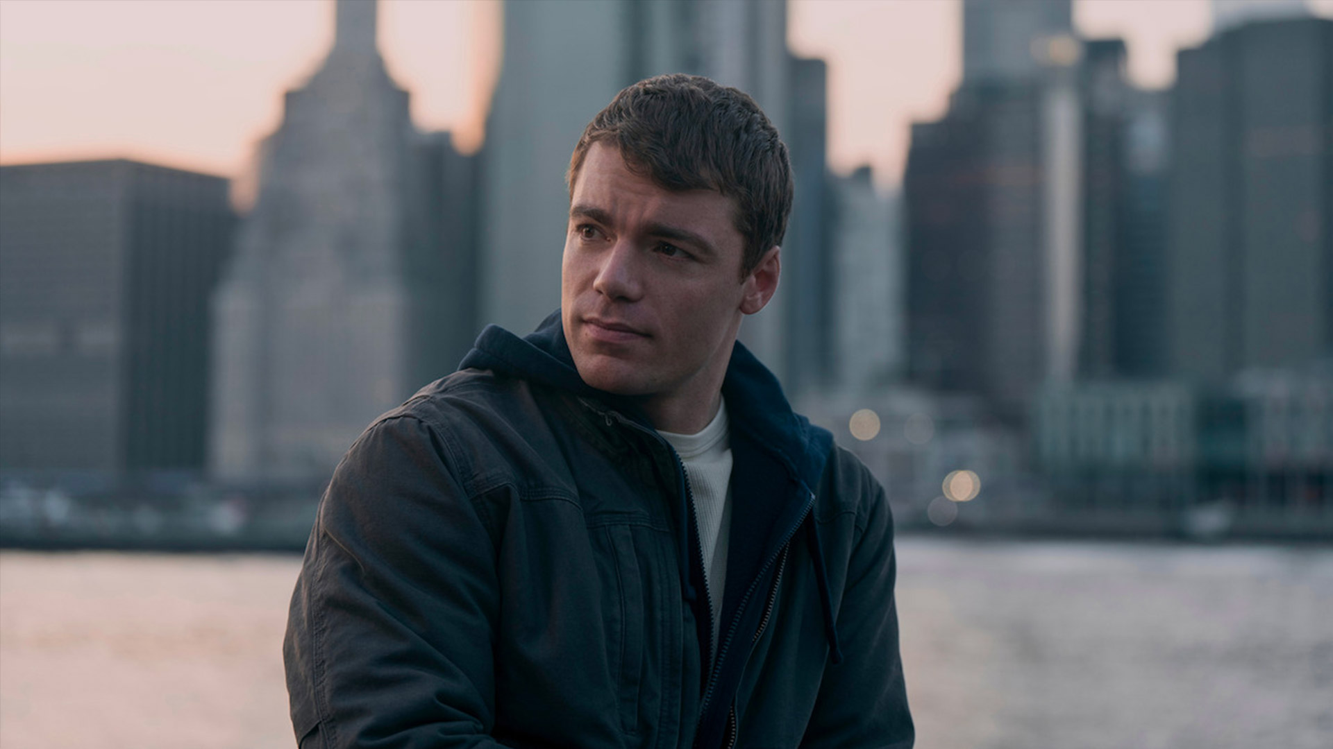 Gabriel Basso looks off camera while standing in front of a city skyline with water in the background 