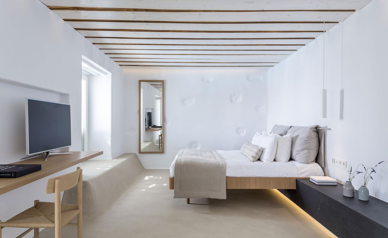 Bedroom in neutral tones with a stone floor, wooden desk and chair and a white ceiling with narrow exposed beams