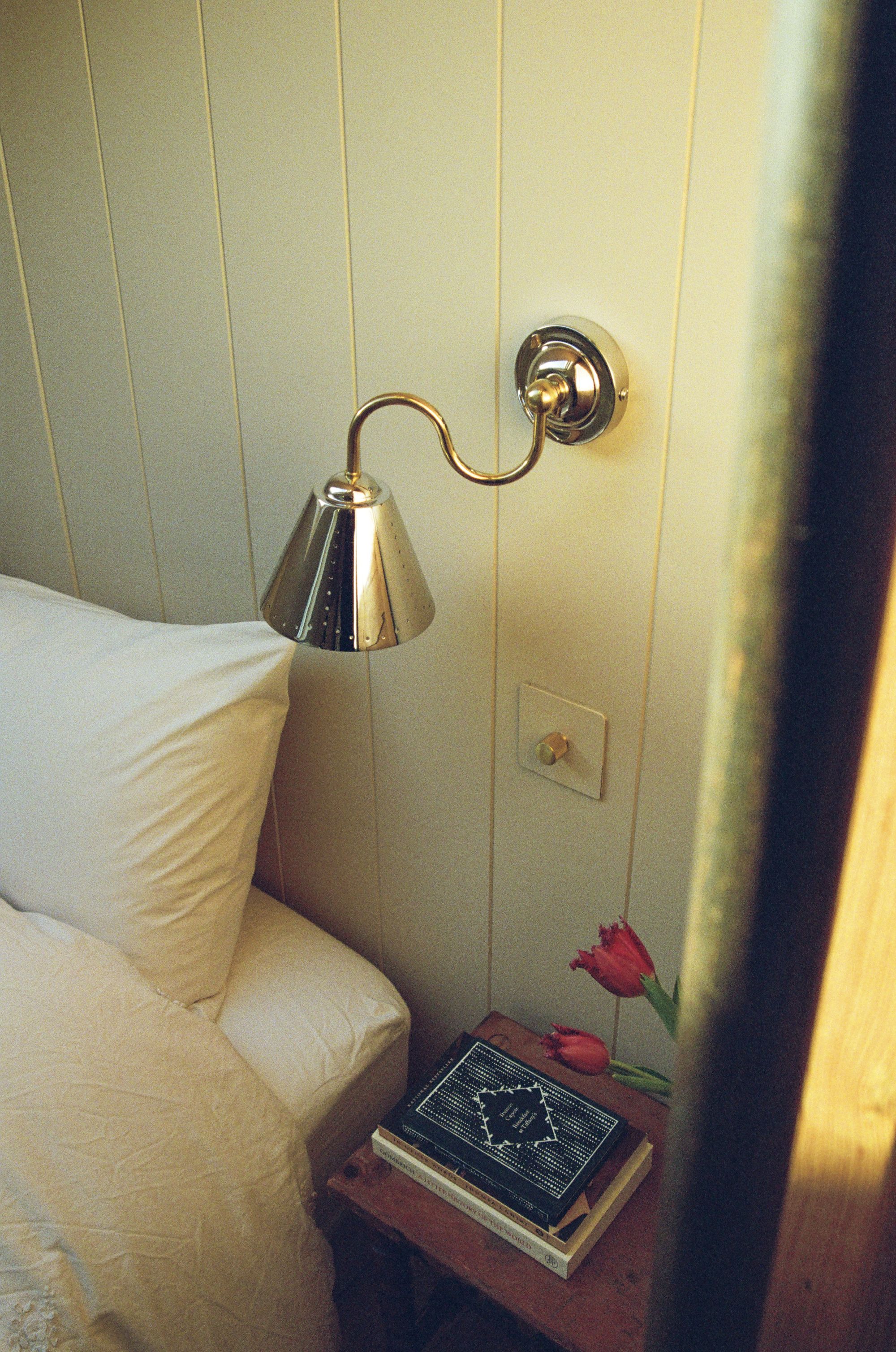 Image of a silver sconce light on a timber wall painted white with the corner of a minimalist but cozy bed in view.