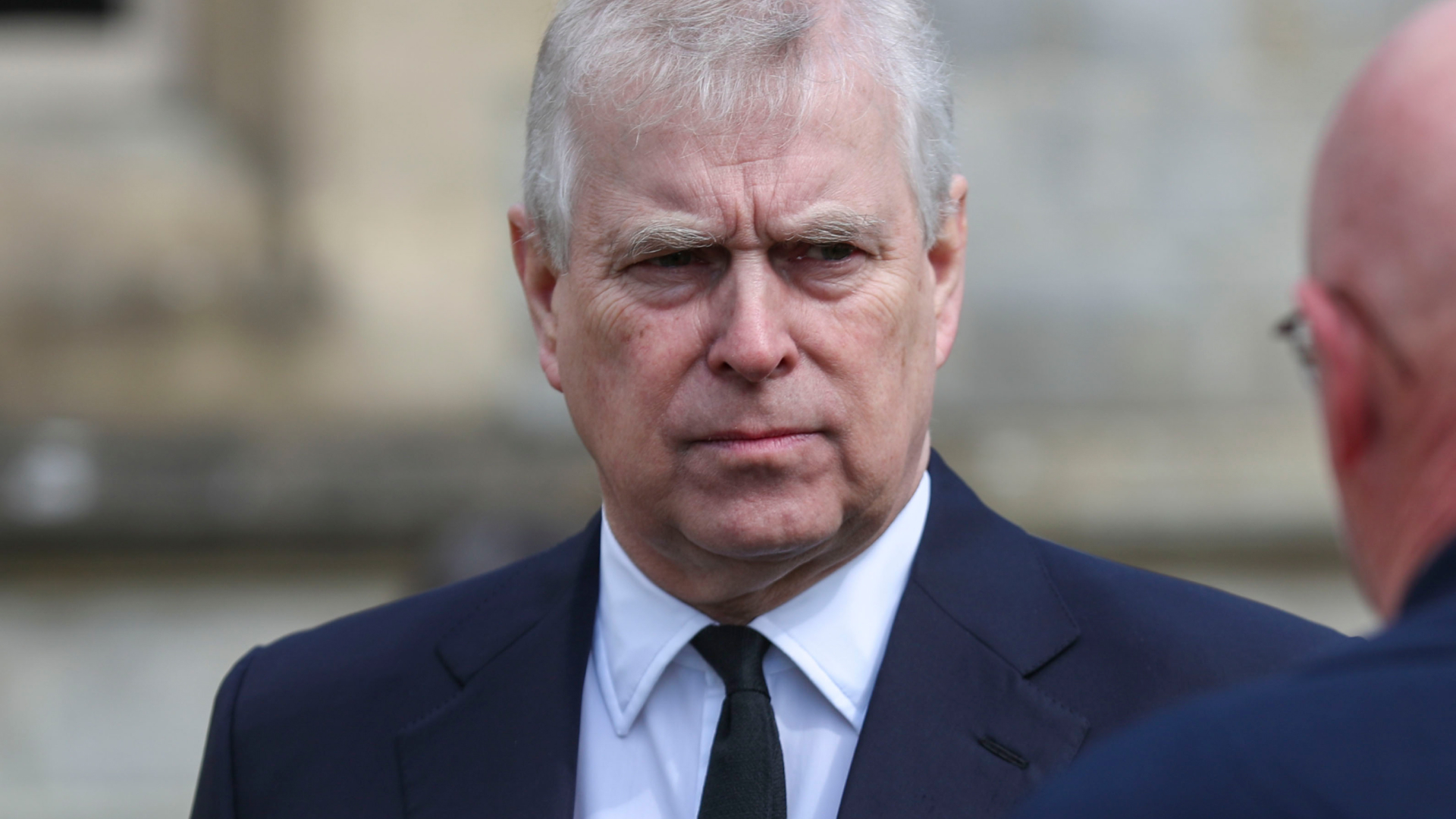 Prince Andrew wears a black suit with a white shirt and black tie and has white gray hair and is scowling