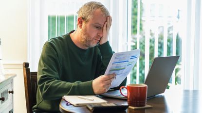 Worried looking man sitting at a table surrounded by documents, his laptop and a calculator