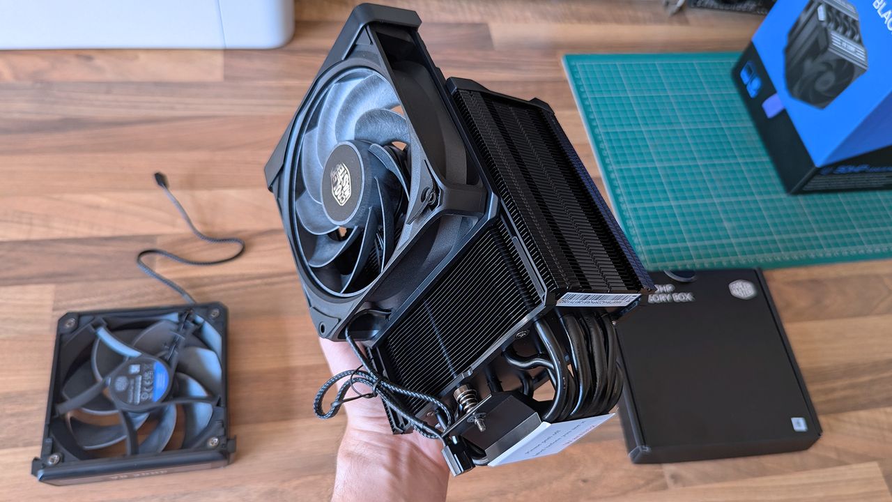 A hand lifts the Cooler Master V8 ACE 3DHP CPU cooler, showing its large heatsink, multiple heat pipes, and mounted fan. Another fan, product box, and PC parts are arranged on the desk below.