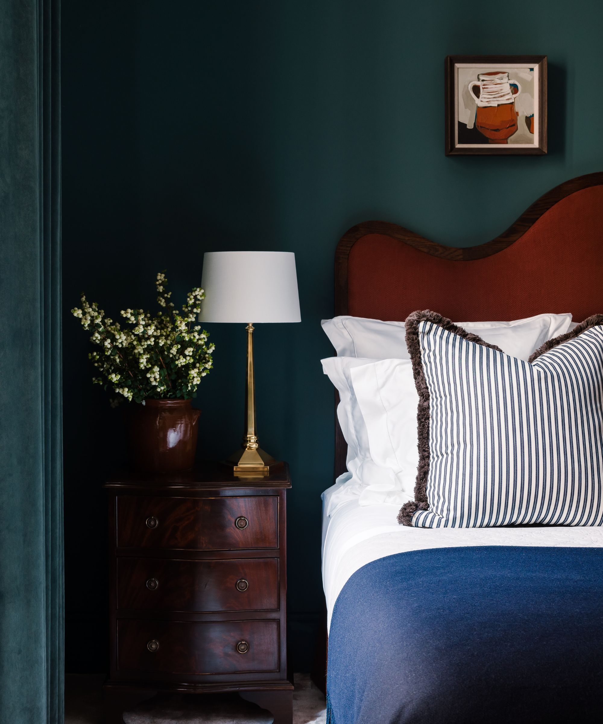 Moody bedroom with dark teal painted walls, an upholstered burnt orange bed, a mahogany wood bedside table, white bedding and a blue throw with a striped cushion in the centre.