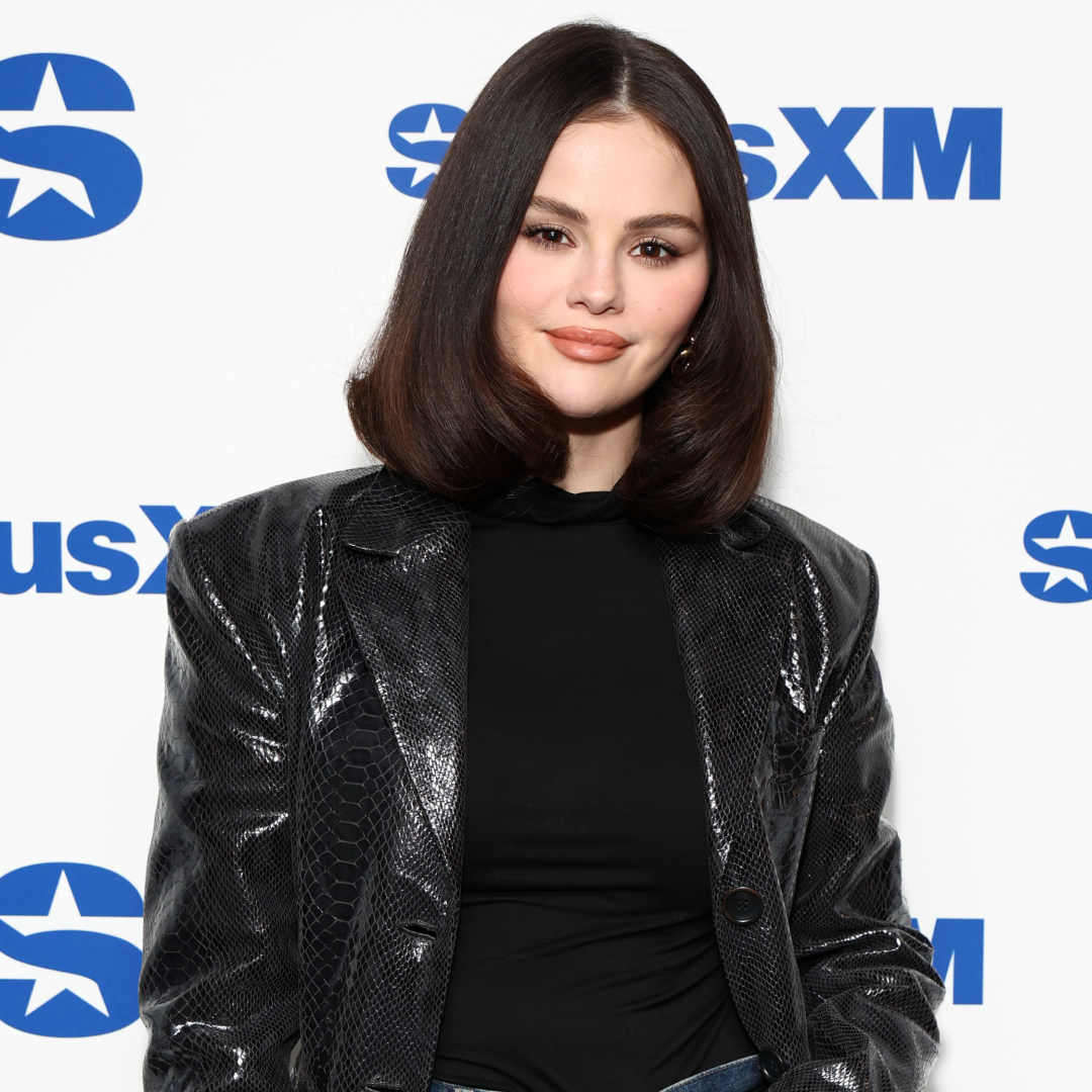 A woman in front of a step and repeat sign with brown hair wearing a leather jacket, soft pink lipstick and soft brown eyeshadow
