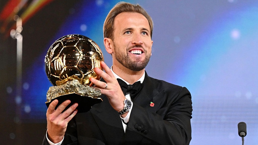 Harry Kane, wearing a black suit and bow tie, lifts the Ballon d'Or trophy.