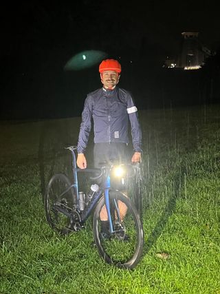 A white man in a blue rain jacket and orange helmet stands in the rain next to a bike