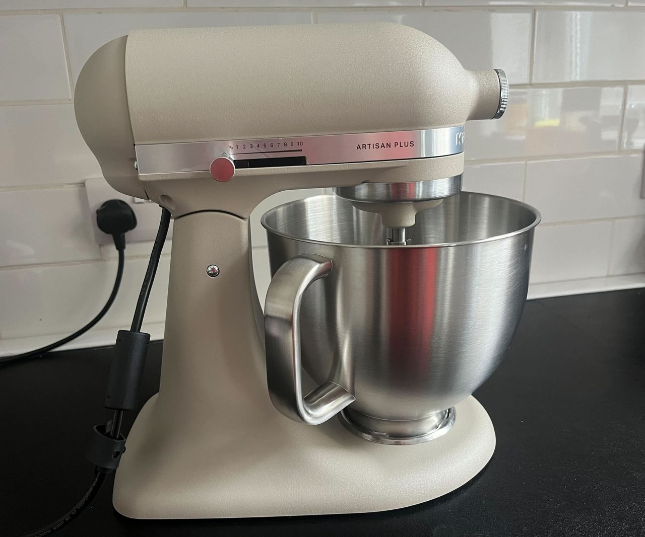 A light beige colored Kitchenaid stand mixer with a stainless steel bowl on a black kitchen counter.