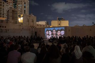 An open courtyard with a cinema style setup. A crowd of people sitting and looking at the image of 5 women projected on the wall. Photographed during the night