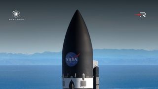 the tip of a black rocket with a nasa meatball logo rises from the center against a hazy blue hilly horizon and greying sky.