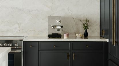 Image of a minimalist kitchen with navy blue cabinetry that has brass hardware and white marble countertops. There is a stylish, concrete espresso machine on the counter next to a pink coffee mug.