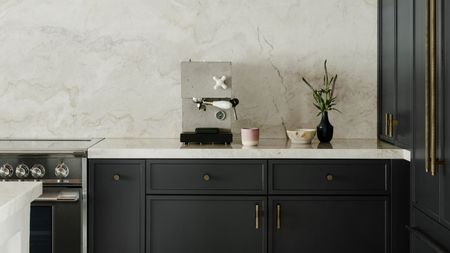 Image of a minimalist kitchen with navy blue cabinetry that has brass hardware and white marble countertops. There is a stylish, concrete espresso machine on the counter next to a pink coffee mug.