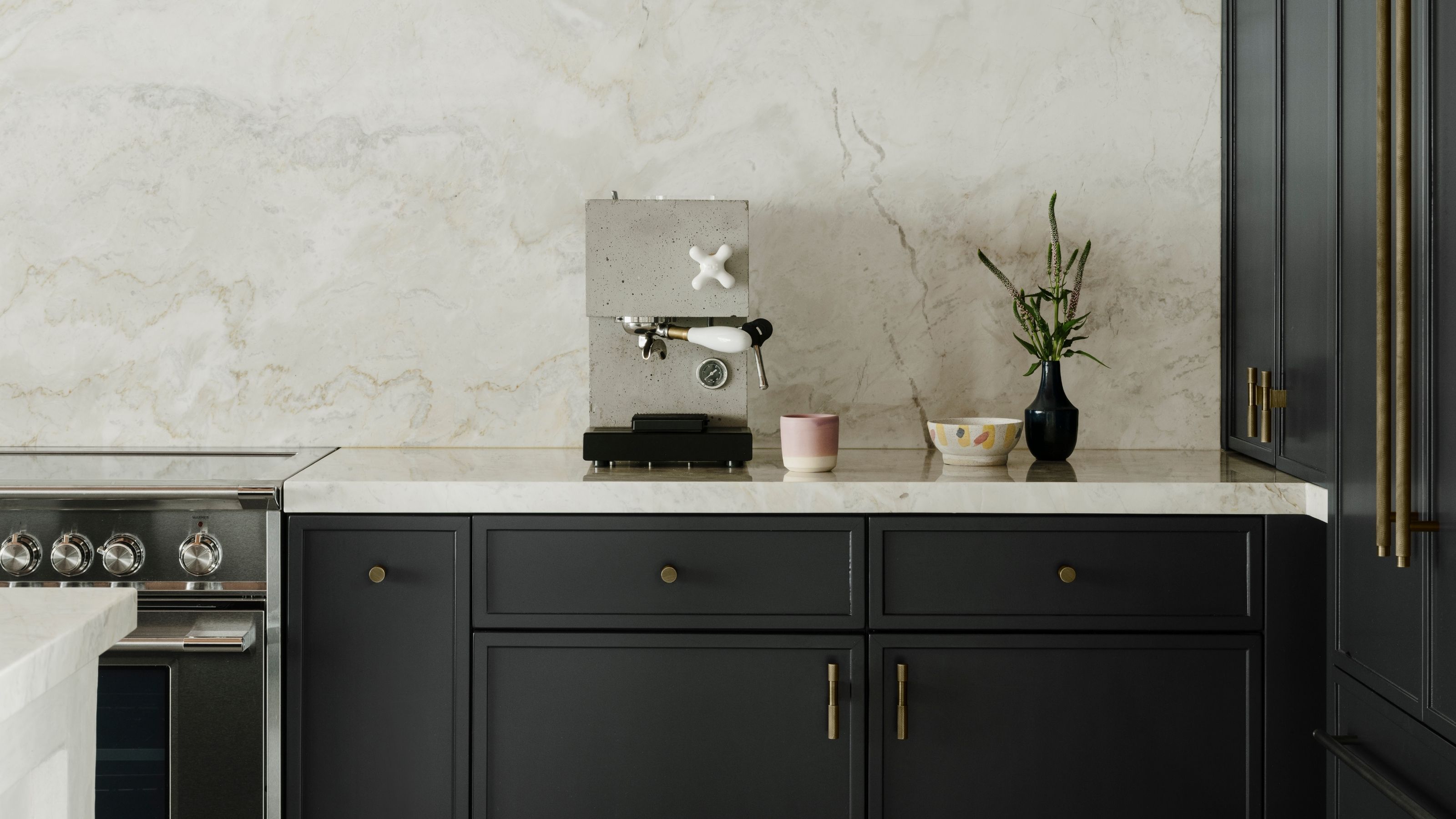 Image of a minimalist kitchen with navy blue cabinetry that has brass hardware and white marble countertops. There is a stylish, concrete espresso machine on the counter next to a pink coffee mug.