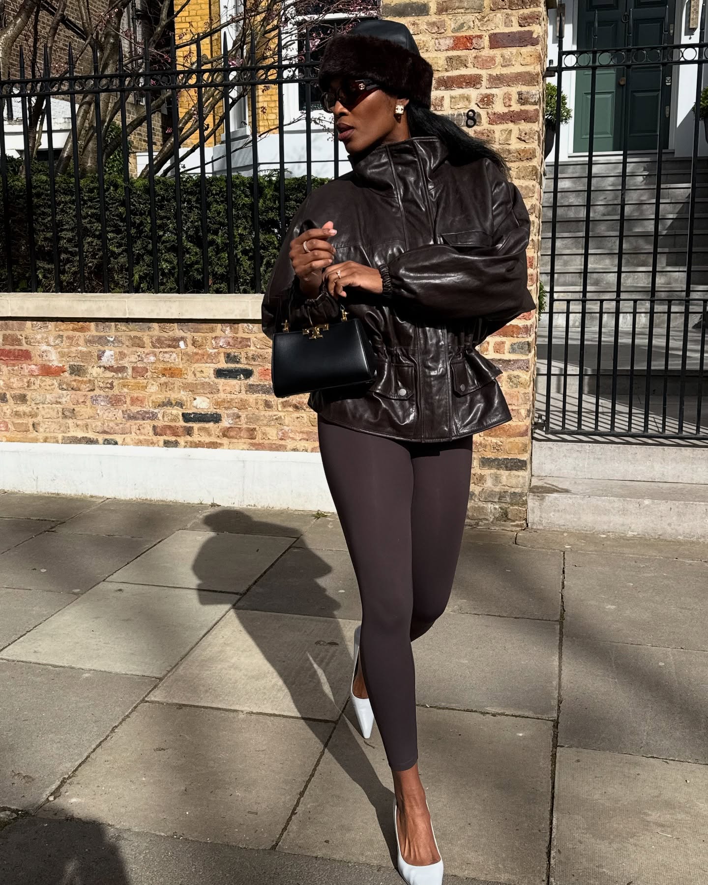 Fashion person nlmarilyn wears a leggings outfit 2026. She wears brown leggings with a cinched waist funnel-neck jacket, a pillbox hat and white curved heels. She finishes the look with a black top handle bag in a recent image taken from her Instagram.
