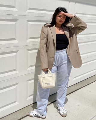 Female mid-size influencer Marina Torres poses in front of a white garage door wearing a neutral blazer, black top, striped pajama pants, and Adidas Samba sneakers.