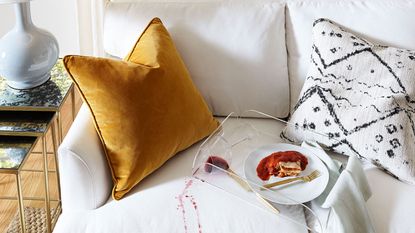 An off-white upholstered sofa with mustard velvet and monochrome textile cushion with red wine spill