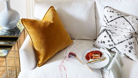 An off-white upholstered sofa with mustard velvet and monochrome textile cushion with red wine spill