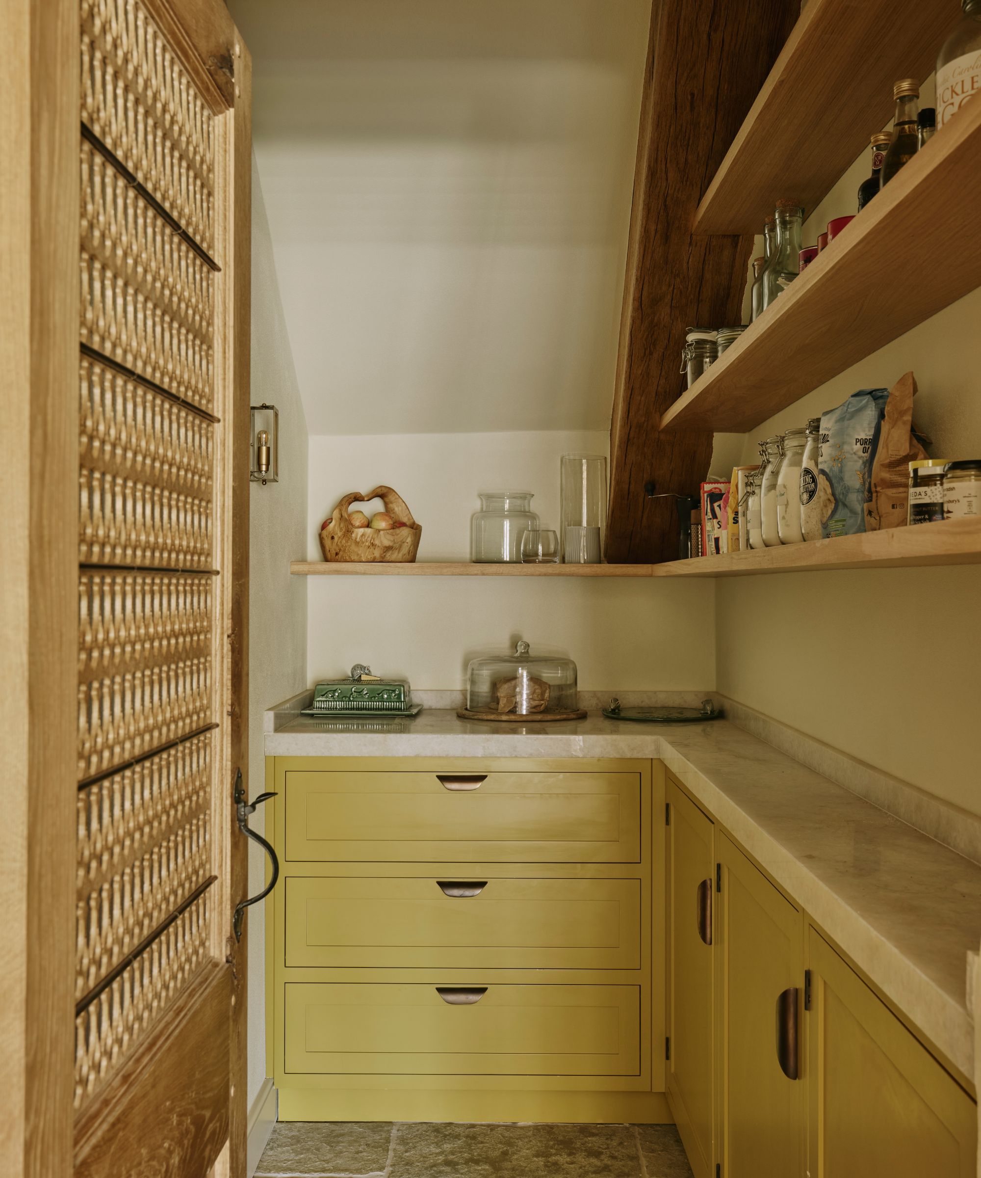 A small walk-in pantry in a Cotswolds kitchen with wooden open shelving, yellow cabinets, and white countertops