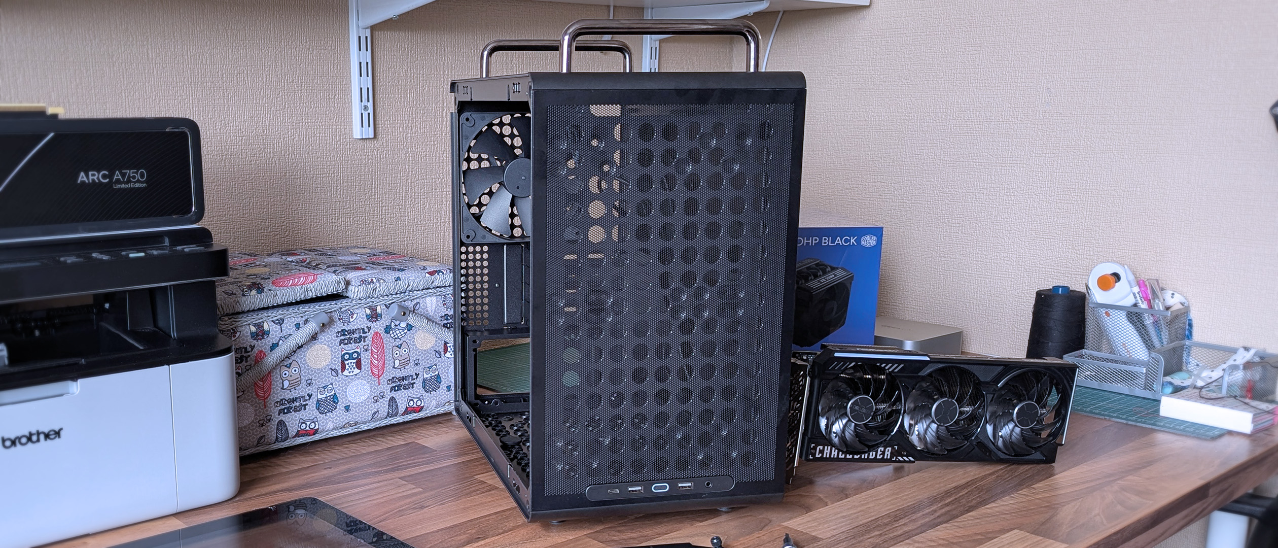 An open Cooler Master Qube 340 PC case sits on a wooden desk, showing its mesh front panel, front I/O ports, and a rear cooling fan inside the chassis.