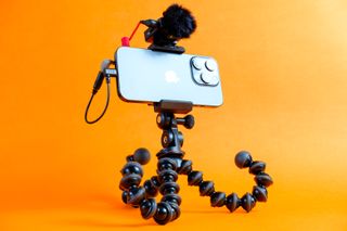 An photo of the Joby GripTight GorillaPod Pro 2 holding an iPhone 15 Pro with a Rode Wireless microphone attached, all set against a vibrant orange background.