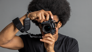 A photographer holding a Fujifilm camera in front of their face, standing in front of a grey background