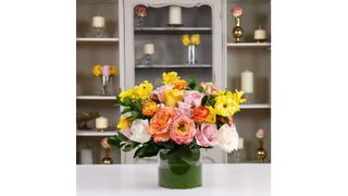 A flower arrangement with yellow, pink, orange, and white roses in a clear vase on a countertop, for the best flower delivery services.