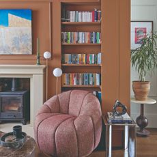 A terracotta-painted living room with a built-in L-shaped bookshelf and a tub accent chair in front of it