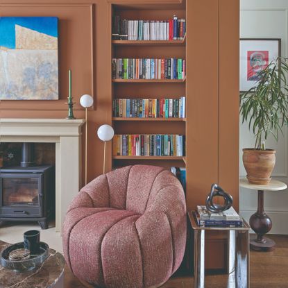 A terracotta-painted living room with a built-in L-shaped bookshelf and a tub accent chair in front of it