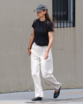 Katie Holmes walking in New York City wearing a baseball cap, black T-shirt, white trousers, and almond-toe black flats.