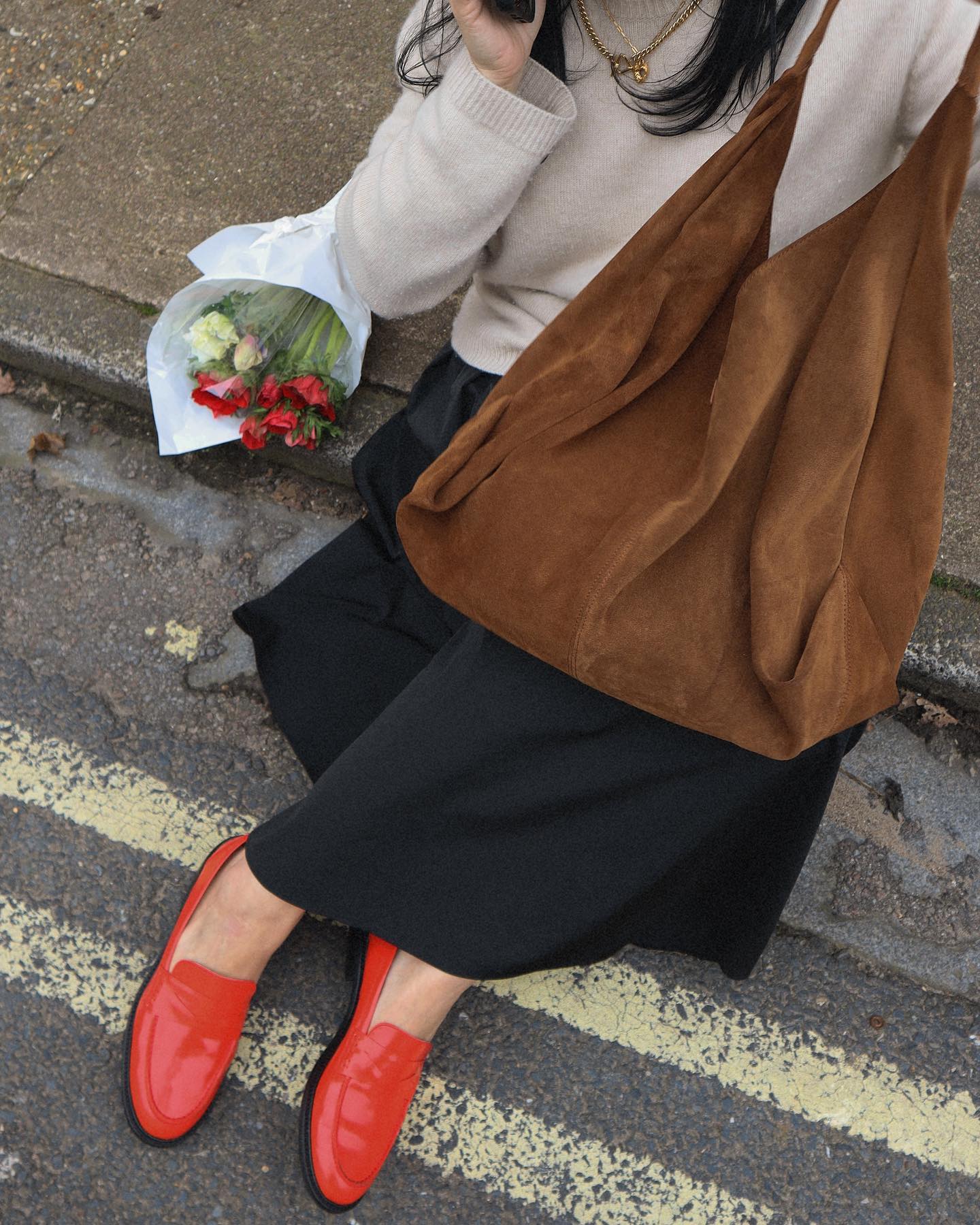 influencer Claudia Berresford sits on a curb next to a bouquet of flowers wearing a neutral sweater, brown suede bag, full black skirt, and red loafers