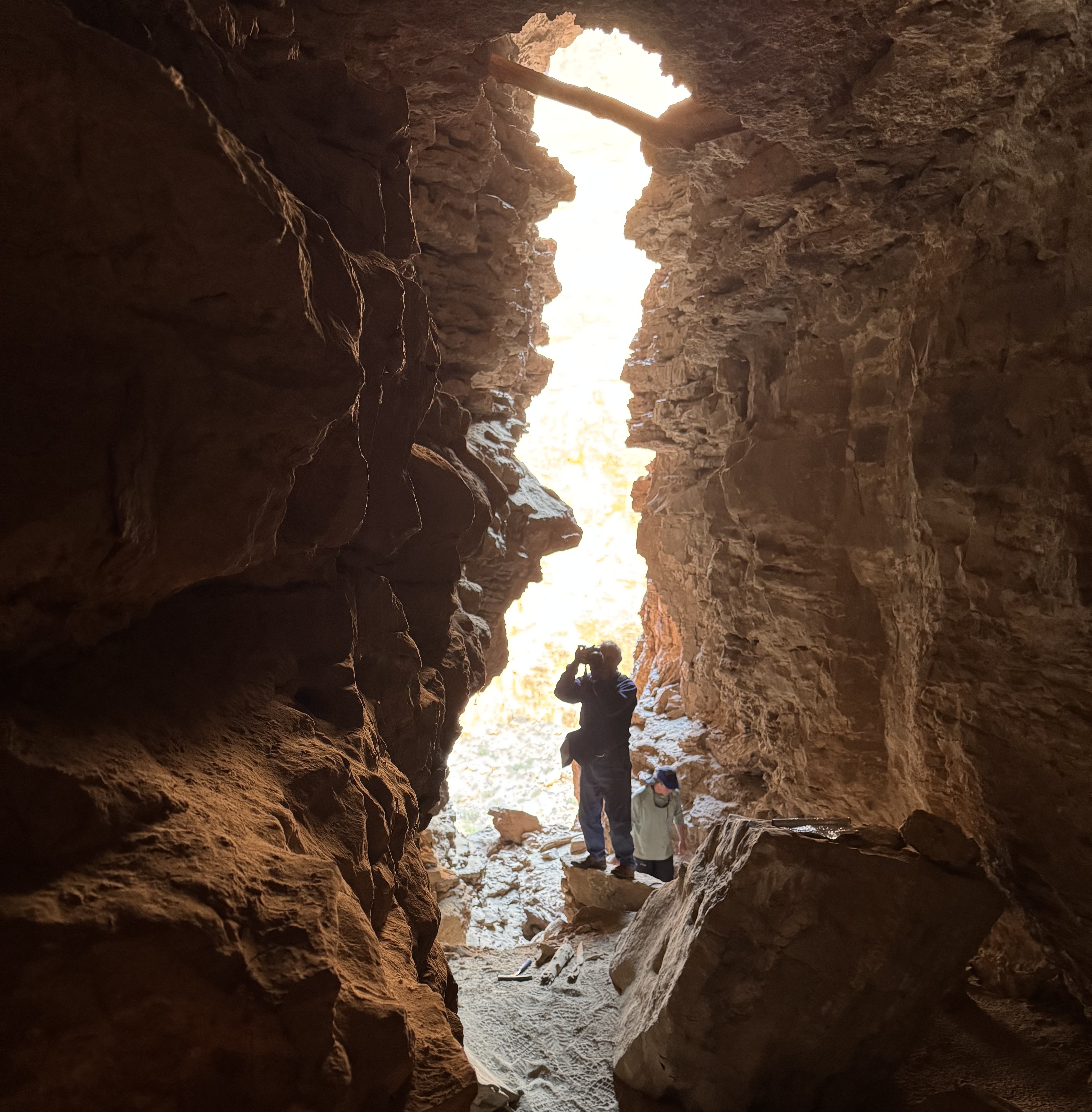 A researchers stands in the entrance of a cave. We see a piece of driftwood stuck between the walls of the cave by its ceiling.
