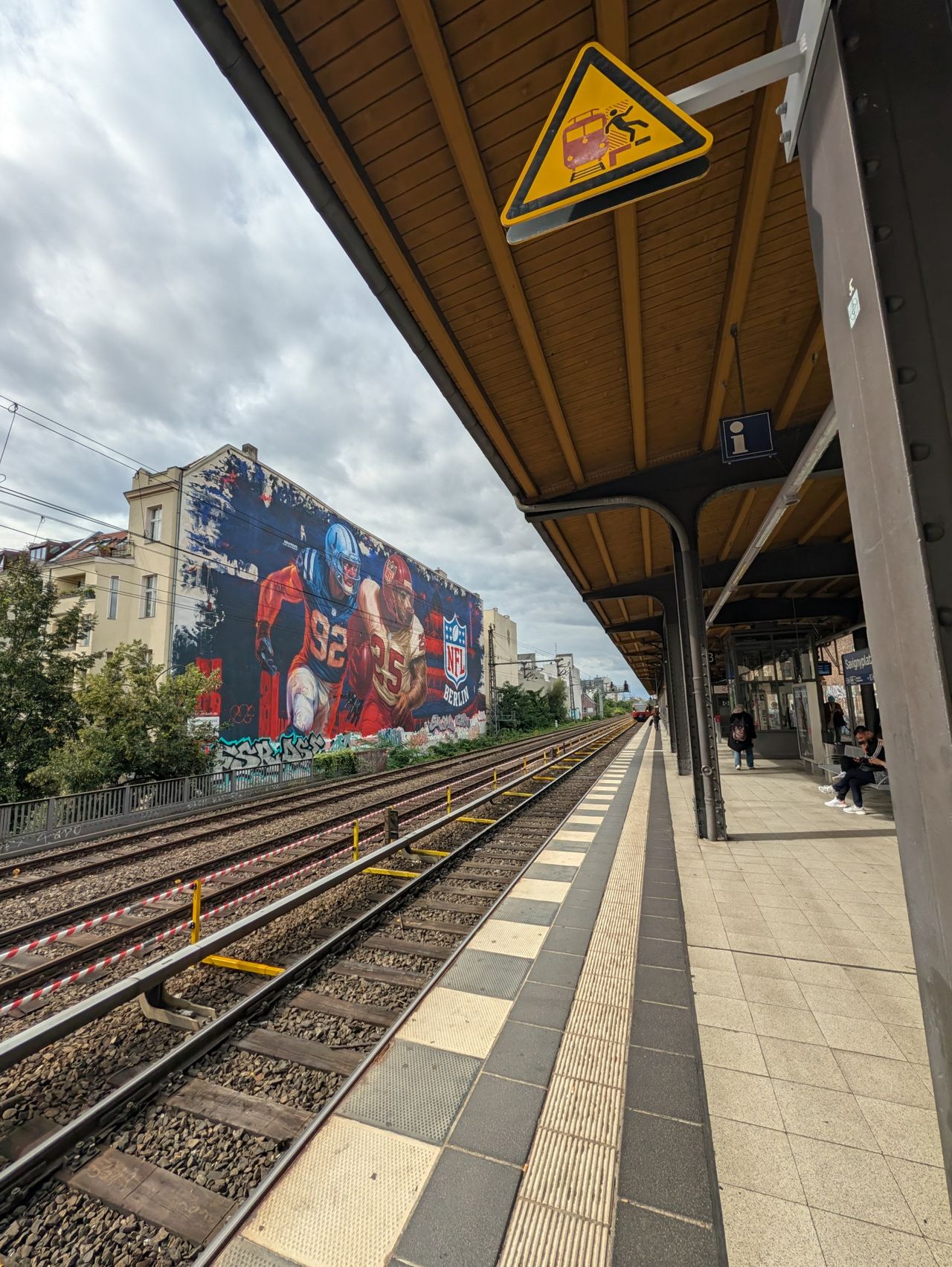 Berlin railway platform with an NFL mural across the tracks and a warning sign overhead.