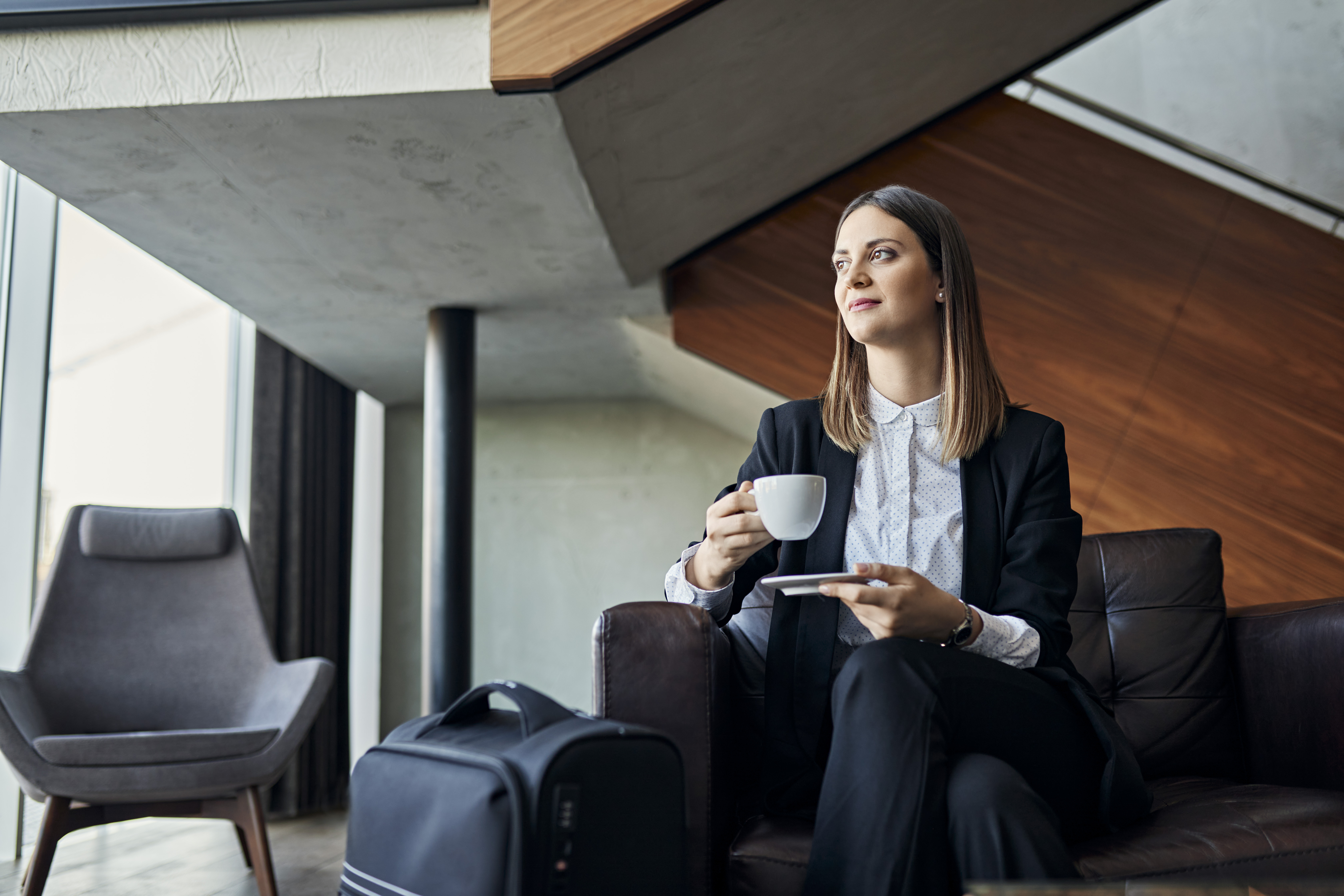 Woman drinks a cup of coffee as she sits in an airport lounge beside her suitcase.