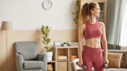 Woman in workout clothes standing in living room with light dumbbell weights in her hands