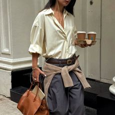 @thecarolinelin wears a pair of grey trousers and a butter yellow shirt with a tan jumper around the waist