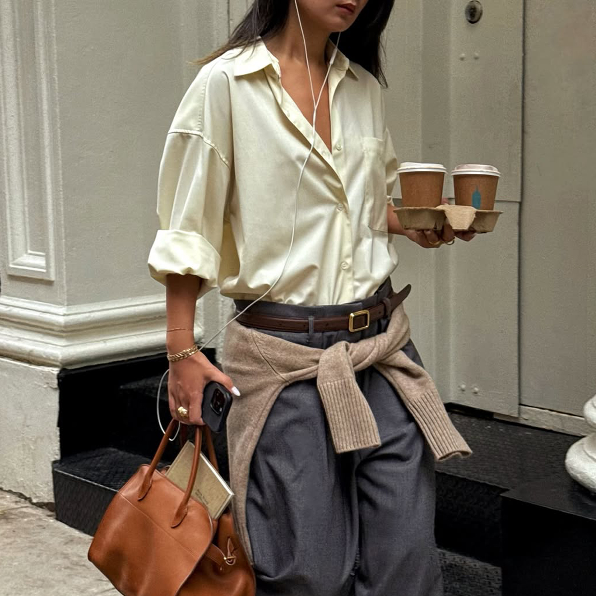 @thecarolinelin wears a pair of grey trousers and a butter yellow shirt with a tan jumper around the waist