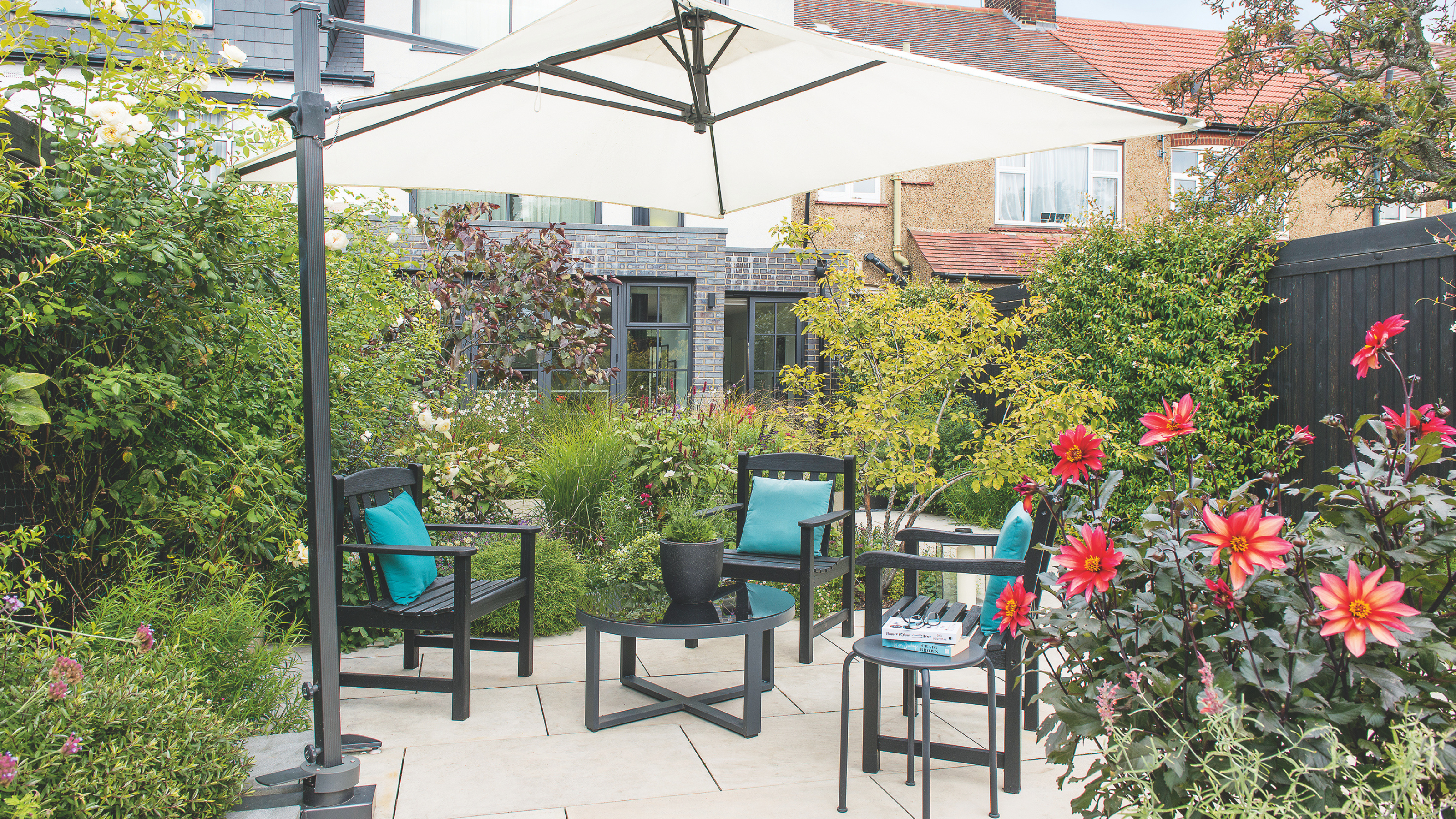 A small garden patio with tables and chairs, a sun umbrella, dense planted flower borders and flowers.