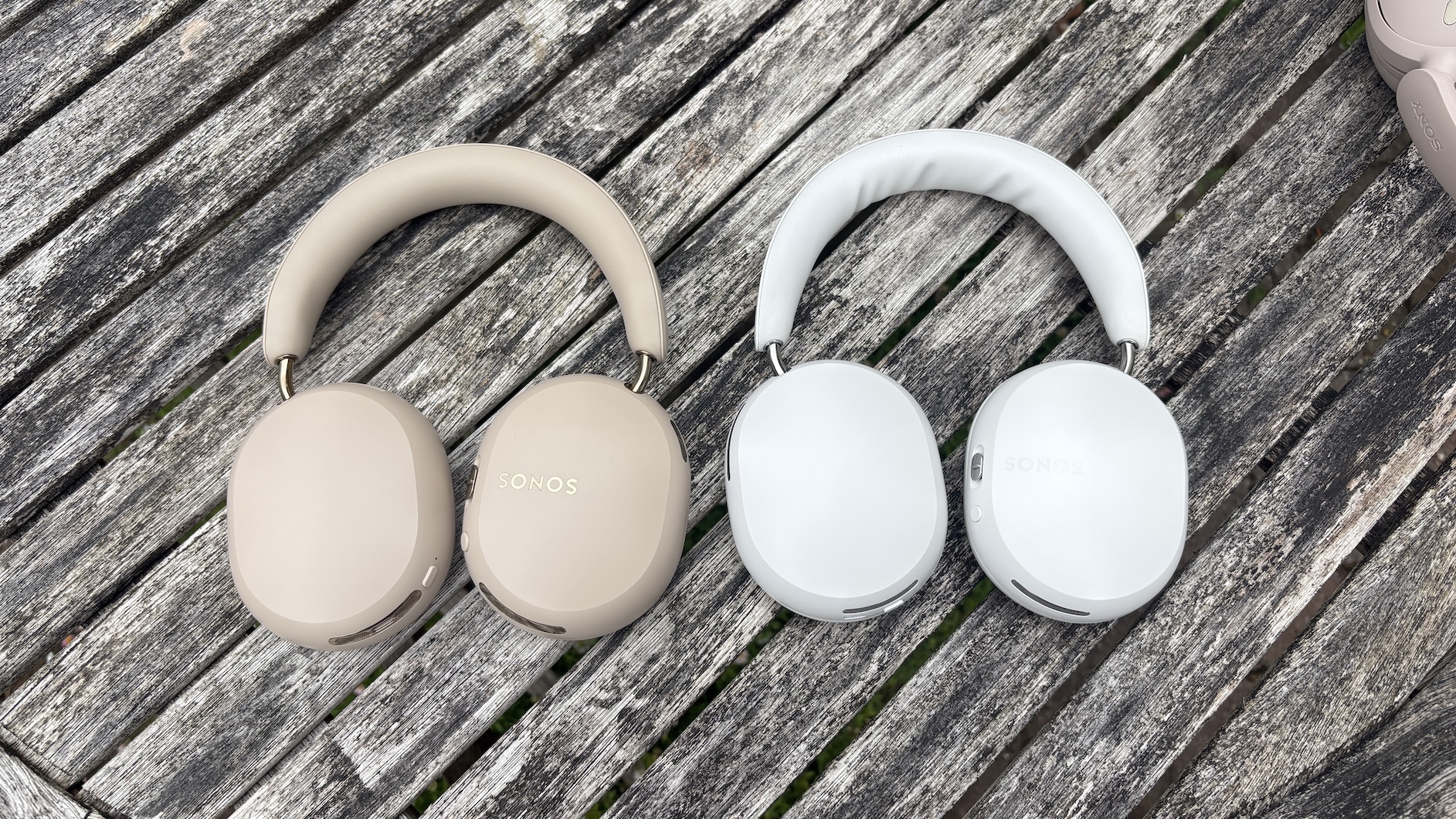 A beige pair of Sonos Ace Ultra headphones next to a white pair of Sonos Ace headphones on a wooden slatted surface, shot from above.