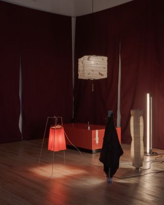 A series of paper lanterns in white, red, black, cream, and decorated patterns stand in a dimly lit room filled with red textiles and furniture.