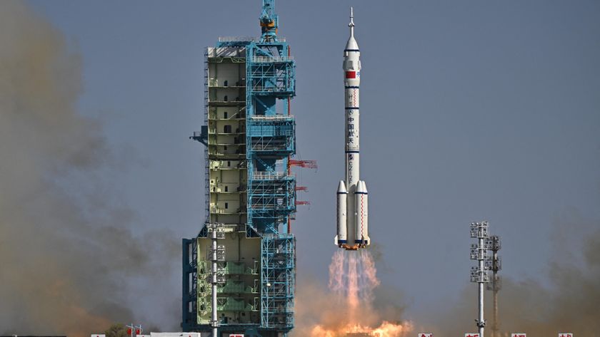 a white rocket with four small boosters at its base lifts off above a plume of fire beside a blue launch tower