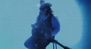 A moody blue shot of Marcus King wearing a cowboy hat, sitting on a seat with his Gibson ES-345