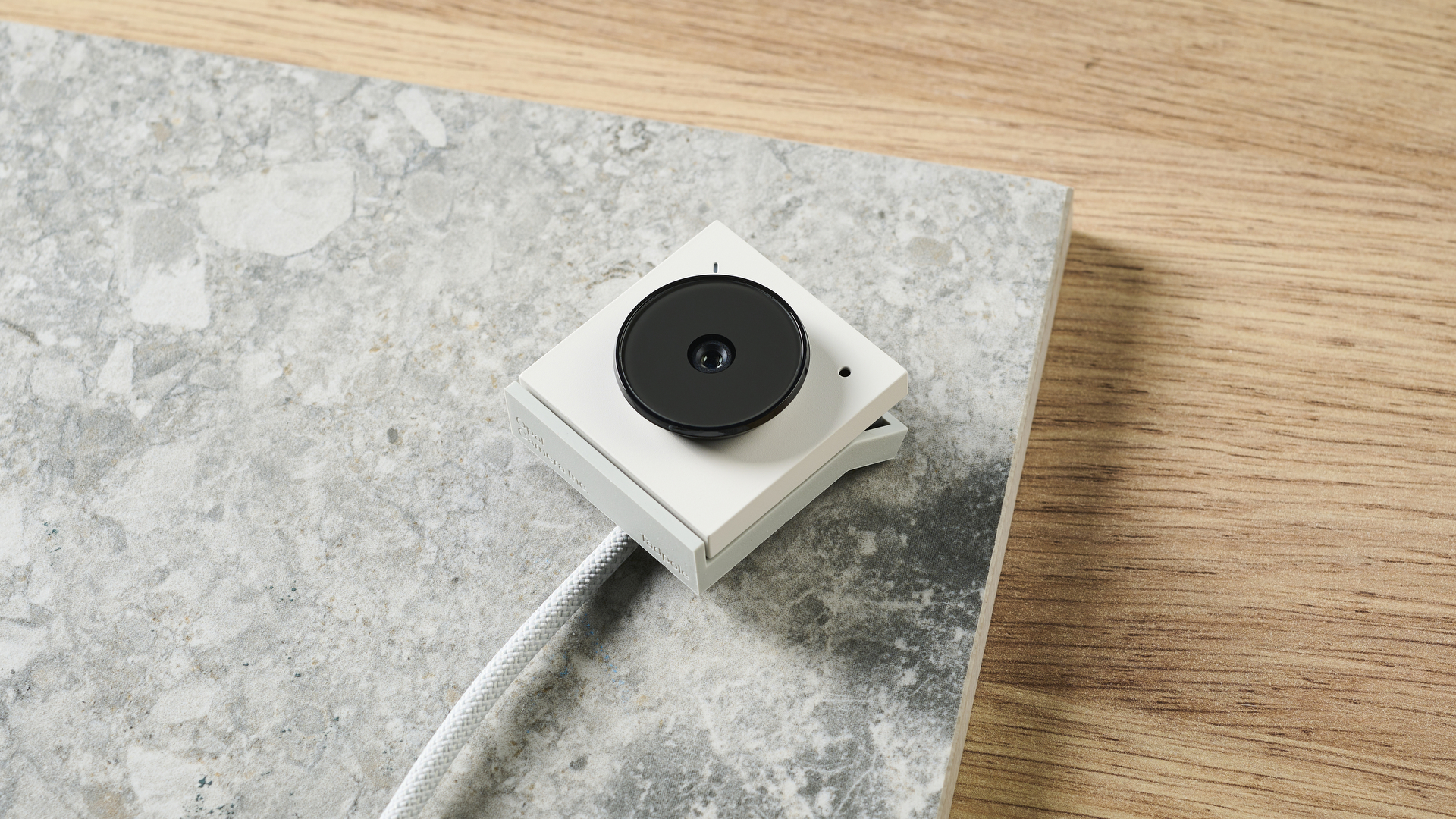 A photo of the Opal Tadpole tiny webcam in white and gray colorway, placed on a stone slate and wooden desk.