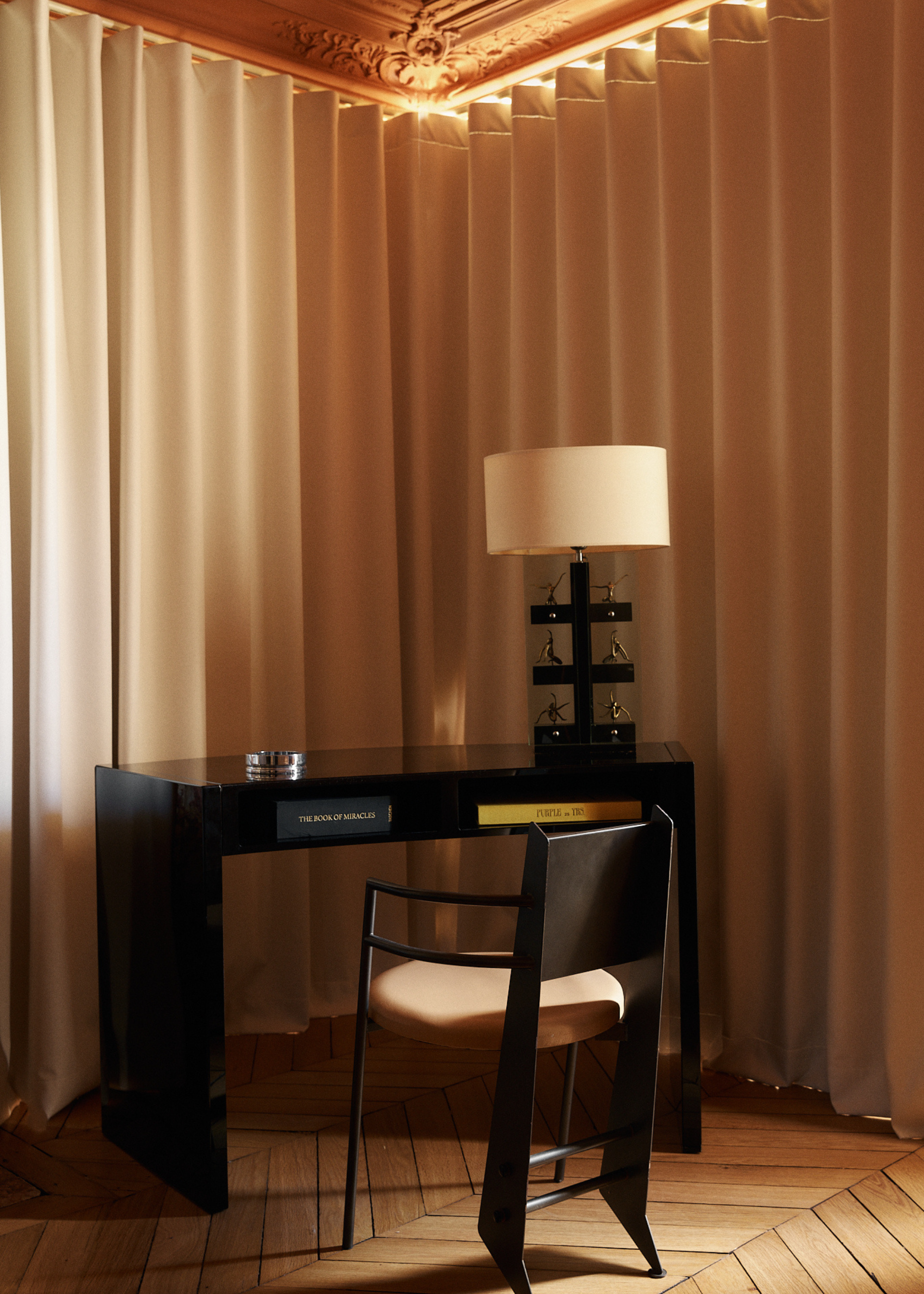 A black desk and chair in the corner of a space surrounded by closed floor-length cream curtains with a black base table lamp with a cream shade