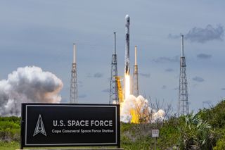 A Falcon 9 rocket lift of from the launch pad. A sign reading "U.S. Space Force Cape Canaveral Space Force Station" is in the foreground of the image and the rocket is launching in the background.